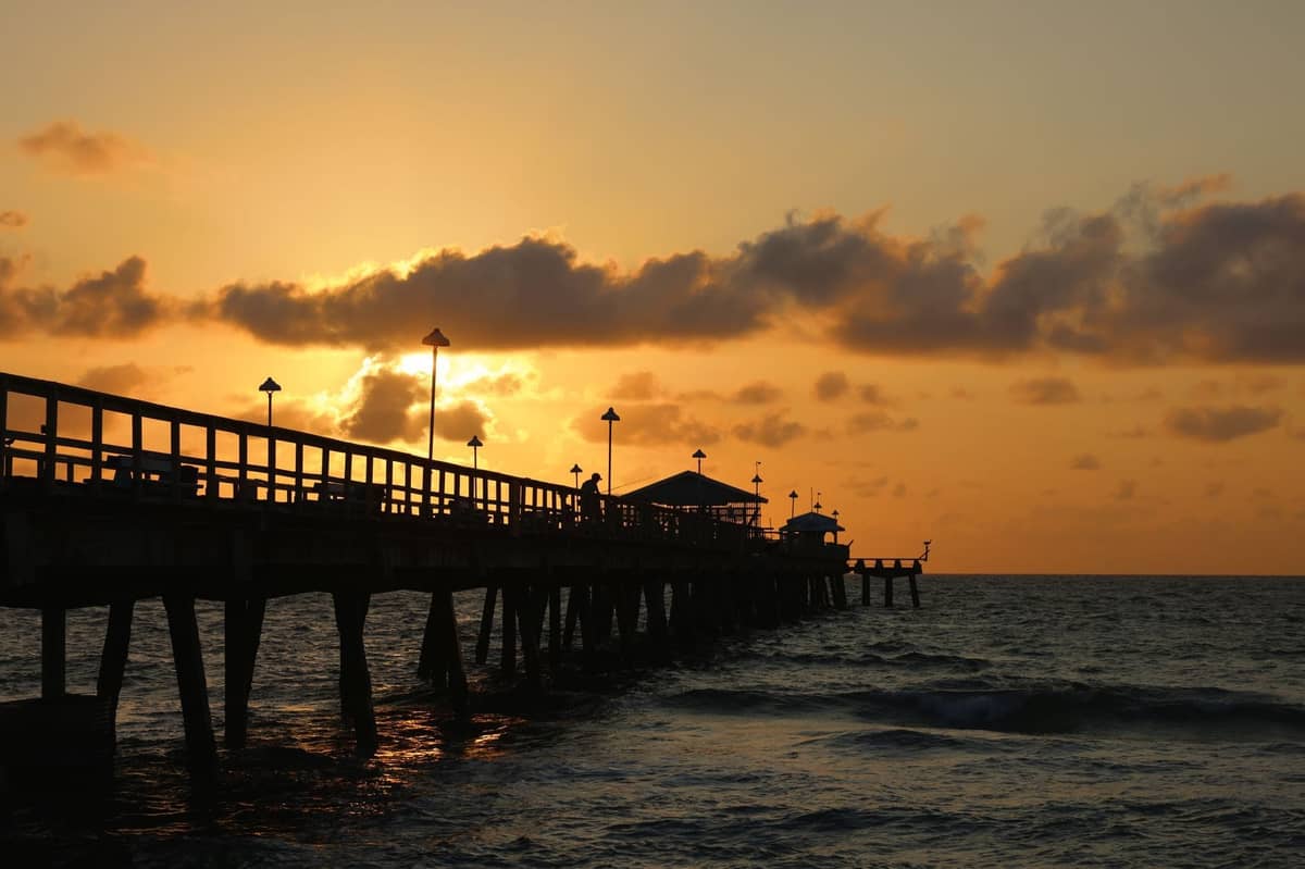 Sunset view of a long fishing pier extending over the ocean, silhouetted against orange sky with scattered clouds.