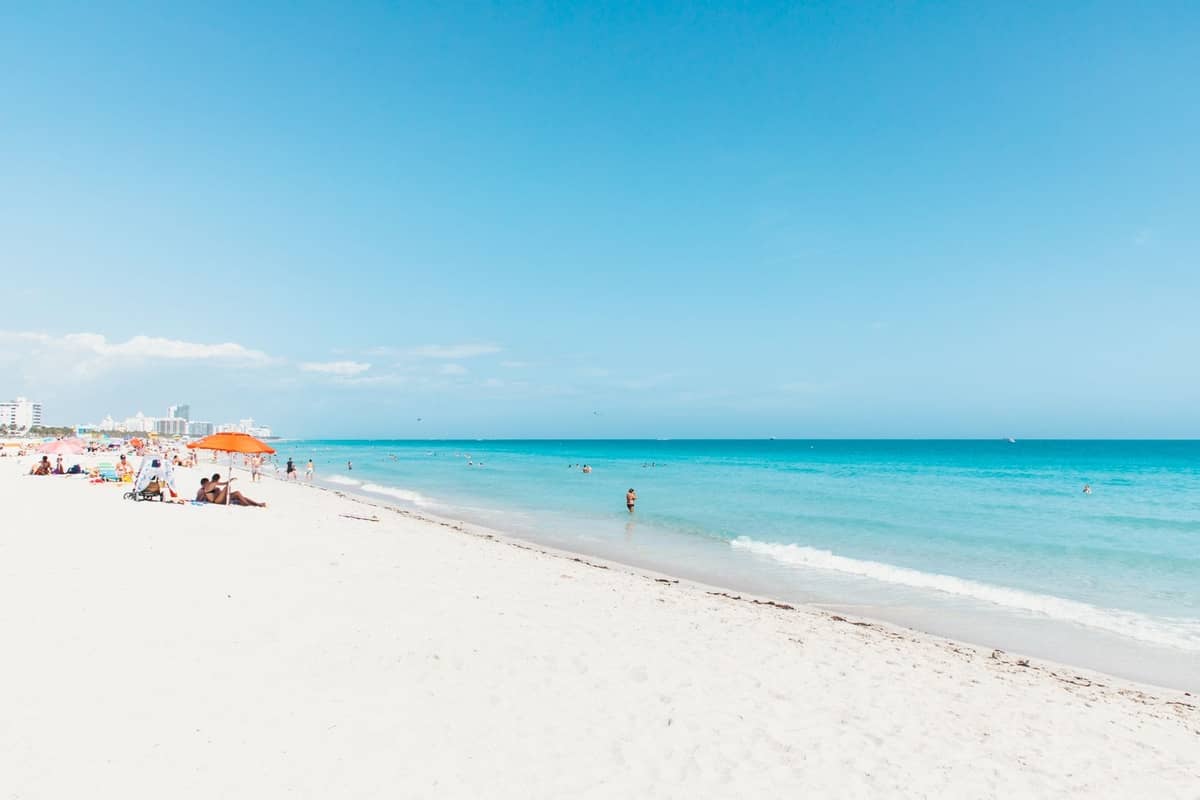 Wide, sunlit view of South Beach in Miami with clear blue sky, turquoise ocean, soft white sand, and people relaxing under umbrellas.