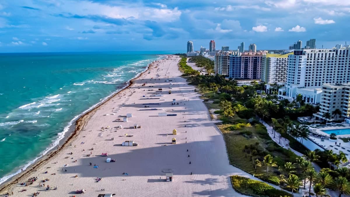 Aerial view of a vibrant Miami beach lined with high-rise hotels and palm trees, featuring wide sand and clear blue water.