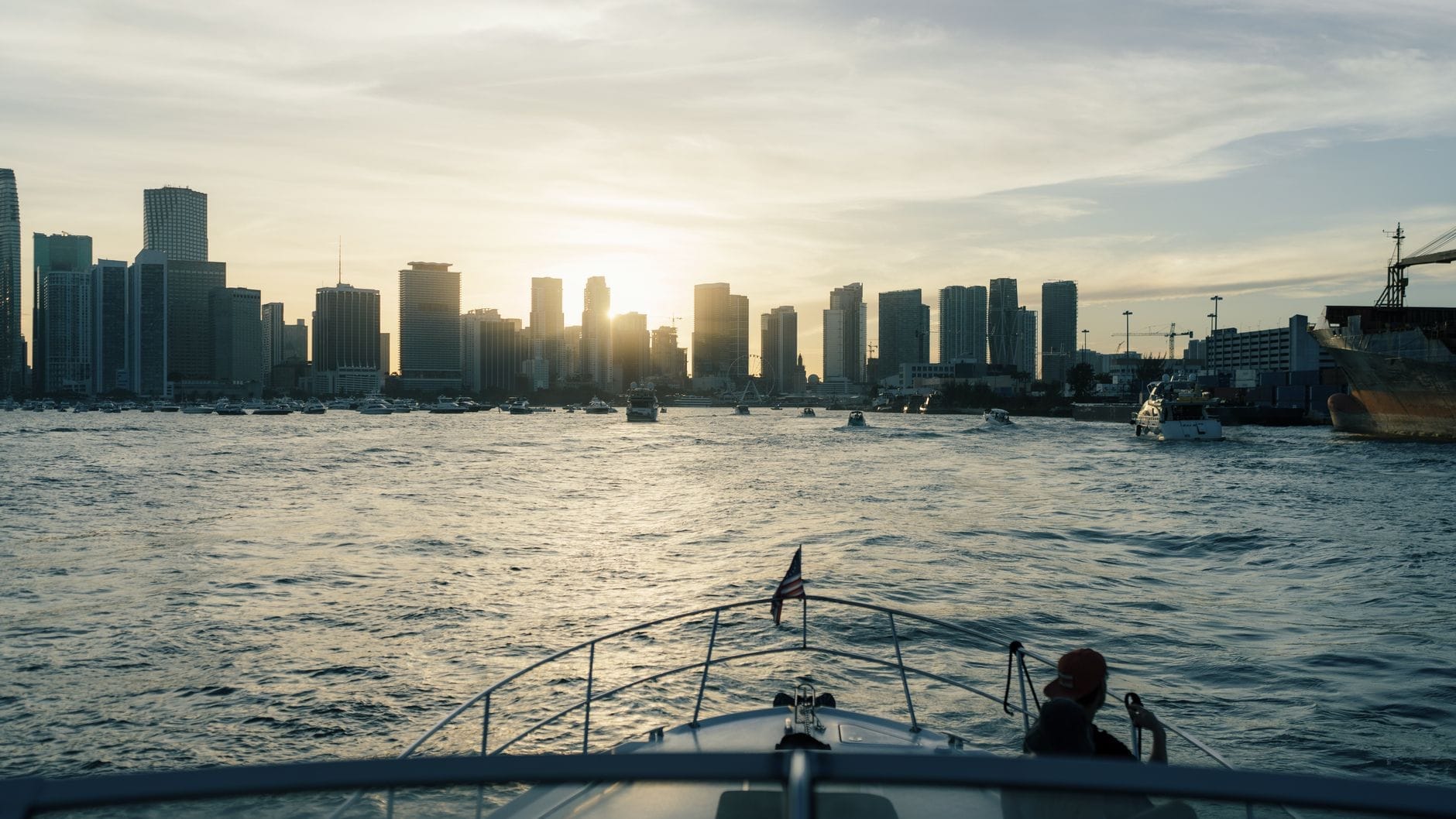 Vista da proa de um barco em direção ao skyline de Miami ao pôr do sol, com outros barcos ao redor na água.