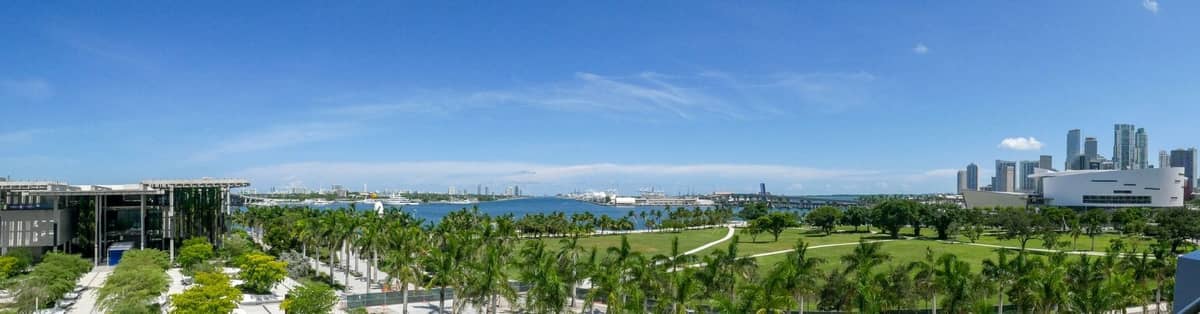 Panoramic view of downtown Miami across Biscayne Bay with lush waterfront park, palm trees, and modern buildings under a blue sky.