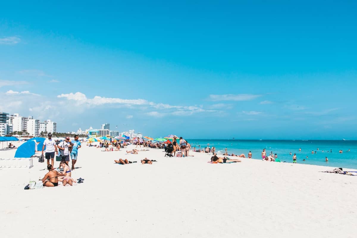 People relaxing on a sunny Miami beach with blue skies, white sand, colorful umbrellas, and buildings in the background.