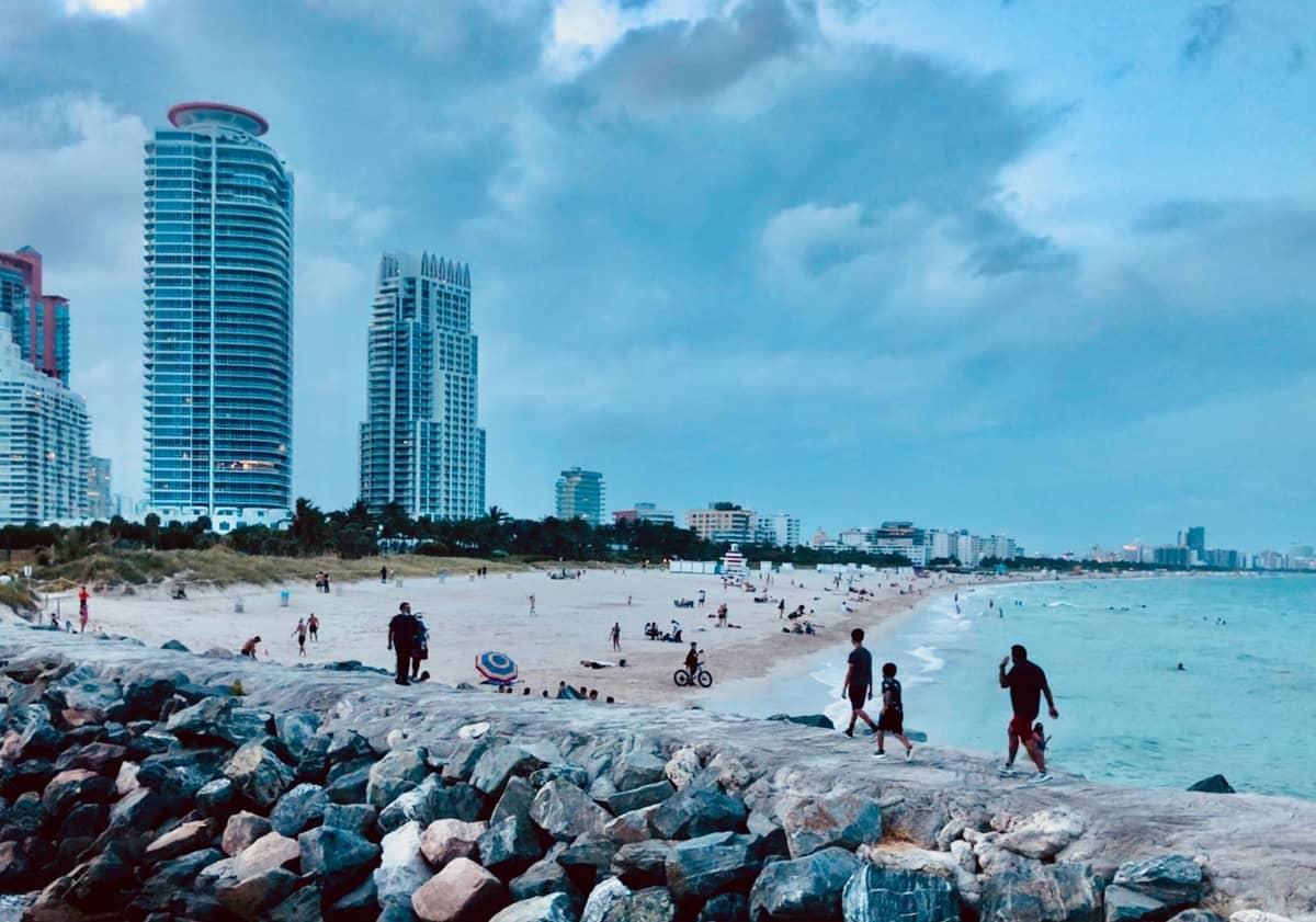 People walking and relaxing on a Miami beach with high-rise buildings in the background and rocks in the foreground.