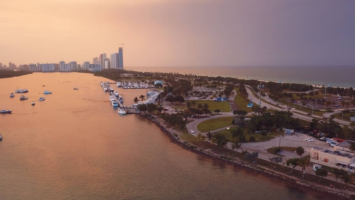 Aerial view of Miami Beach Marina at sunset with yachts docked, road access, and Miami Beach skyline in the background.