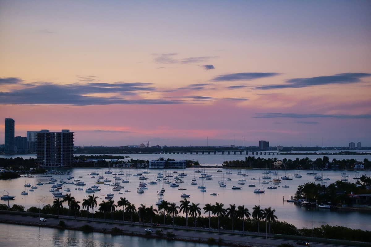 Dozens of boats anchored in Biscayne Bay at sunset, with palm trees, Miami skyline, and bridges visible in the background.