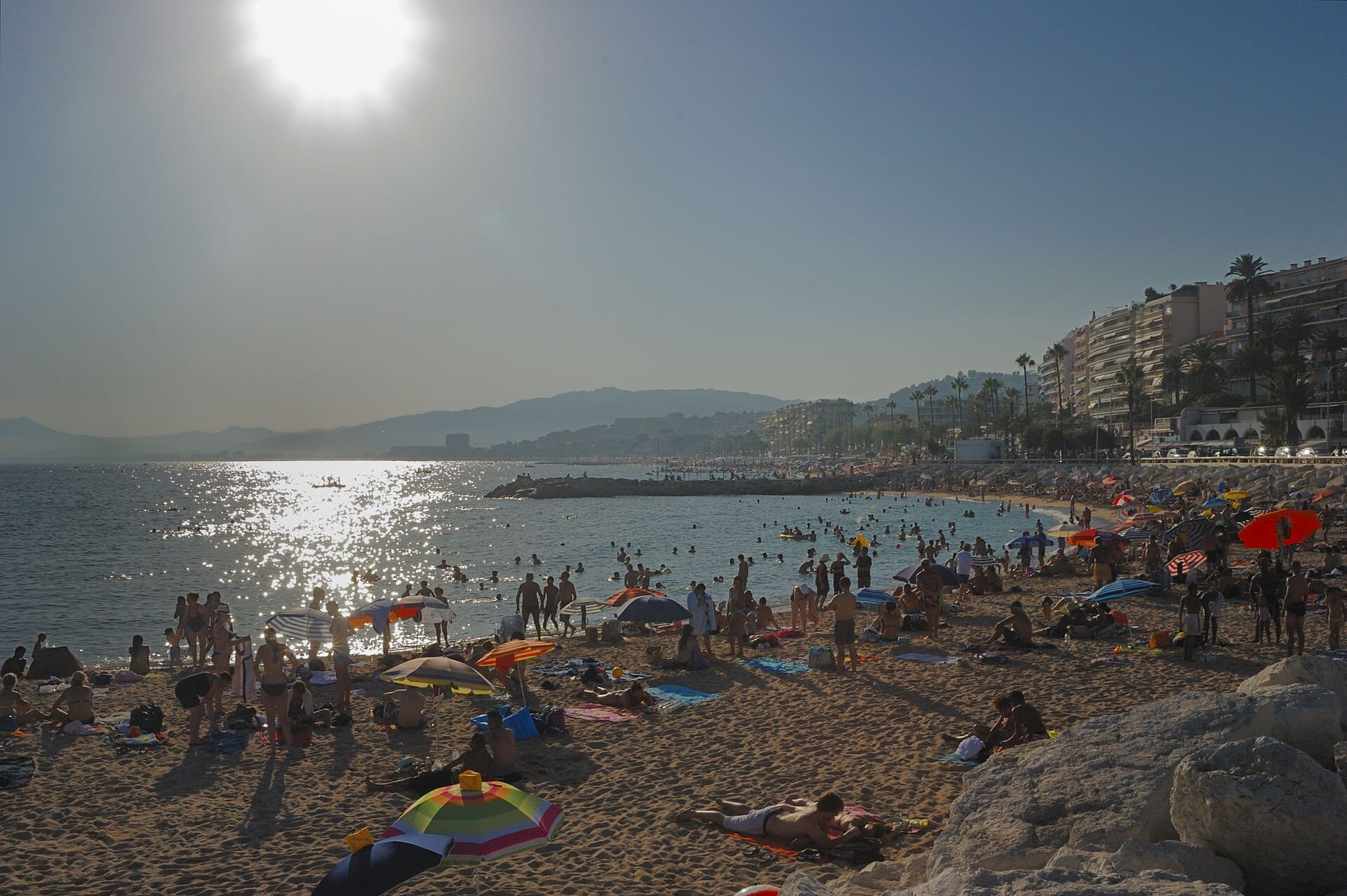 Affollata Plage du Midi a Cannes con bagnanti, ombrelloni colorati, acqua scintillante e palazzi di appartamenti lungo la riva soleggiata.