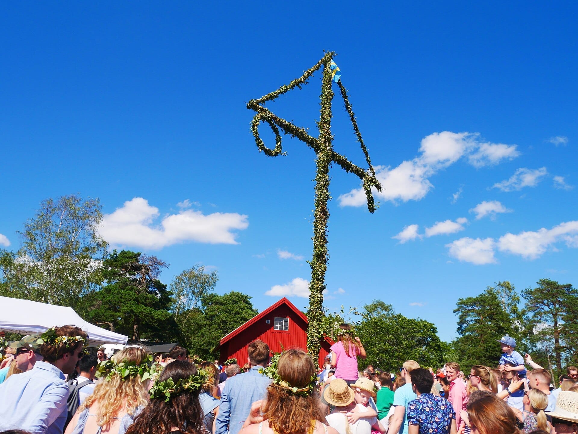 ฝูงชนสวมมงกุฎดอกไม้รวมตัวกันรอบเสา Maypole ที่ประดับตกแต่งที่ Skansen ในสตอกโฮล์มระหว่างเทศกาล Midsommar ใต้ท้องฟ้าสีฟ้าสดใส