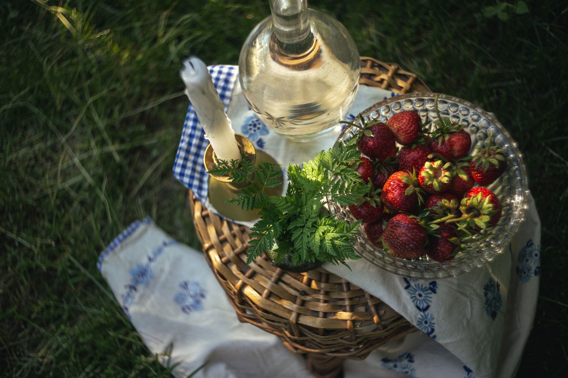 การจัดโต๊ะ Midsommar แบบสวีเดนกลางแจ้ง พร้อมชามแก้วที่เต็มไปด้วยสตรอว์เบอร์รีสด เหยือกน้ำ และพืชสีเขียวบนถาดสานที่วางบนผ้าสีฟ้าและขาว
