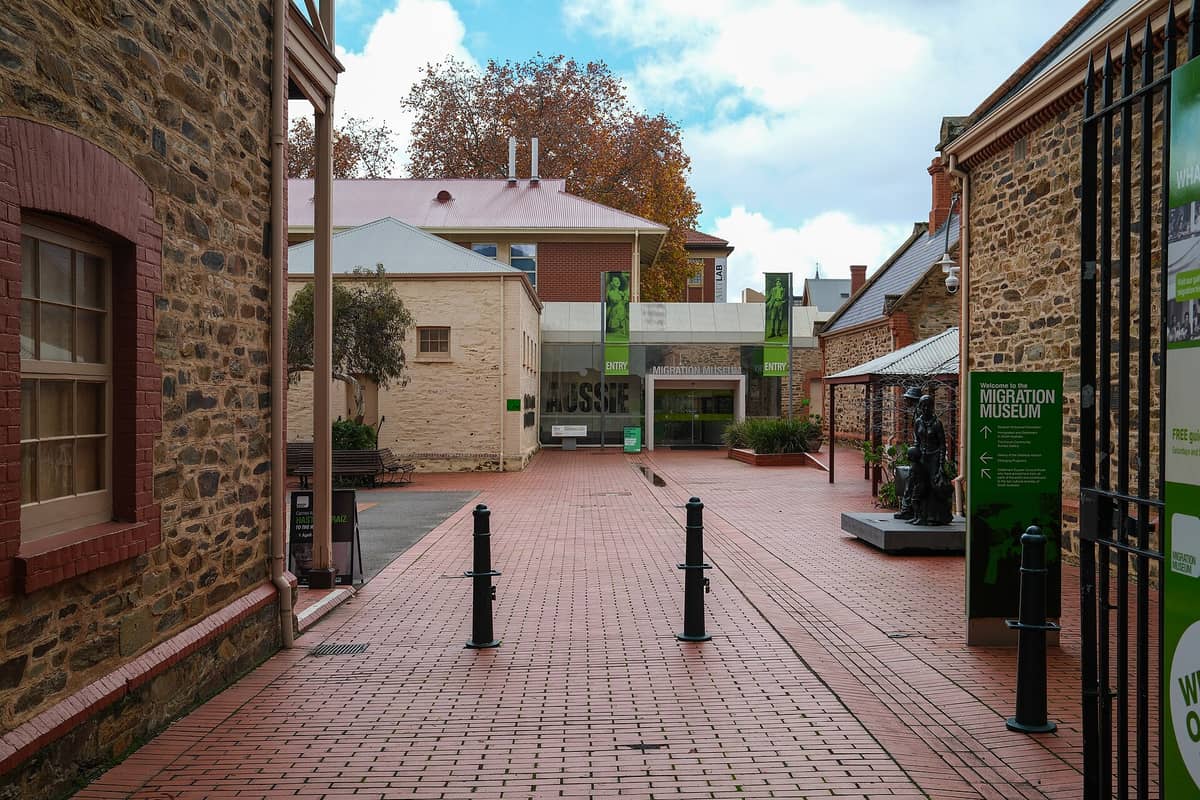 Entrance courtyard to the Adelaide Migration Museum with brick paths, historic stone buildings, a welcome sign, and the museum entryway visible ahead.