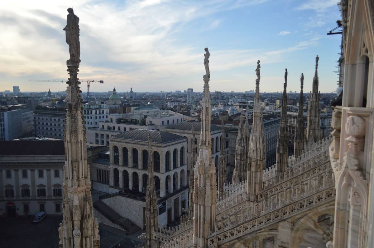 Widok na panoramę Mediolanu z tarasów dachowych Duomo, ukazujący ozdobne iglice i pejzaż miejski pod jasnym niebem.