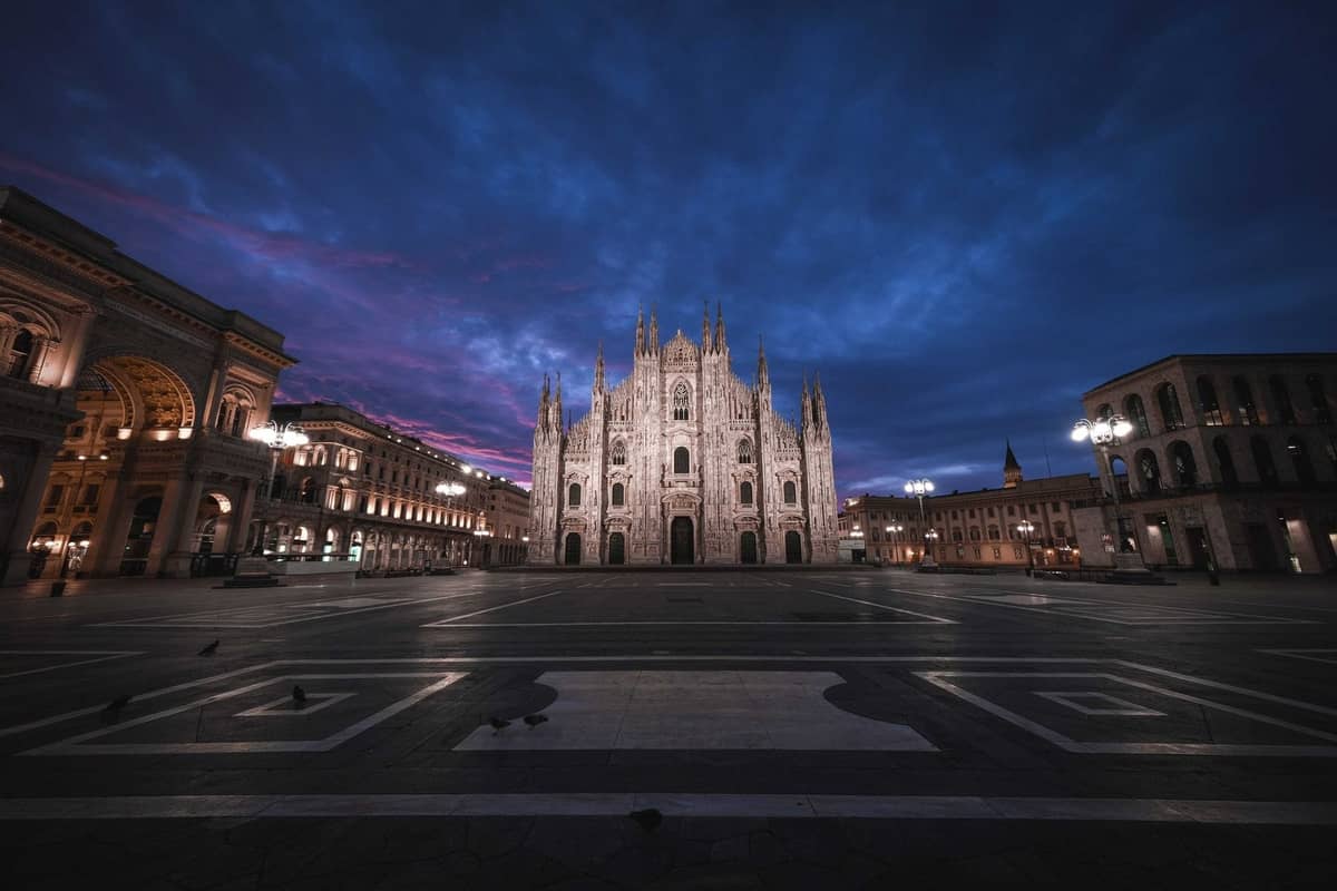 Szeroki widok na Katedrę w Mediolanie (Duomo di Milano) i otaczający plac o świcie, z dramatycznym błękitnym niebem i podświetloną fasadą.