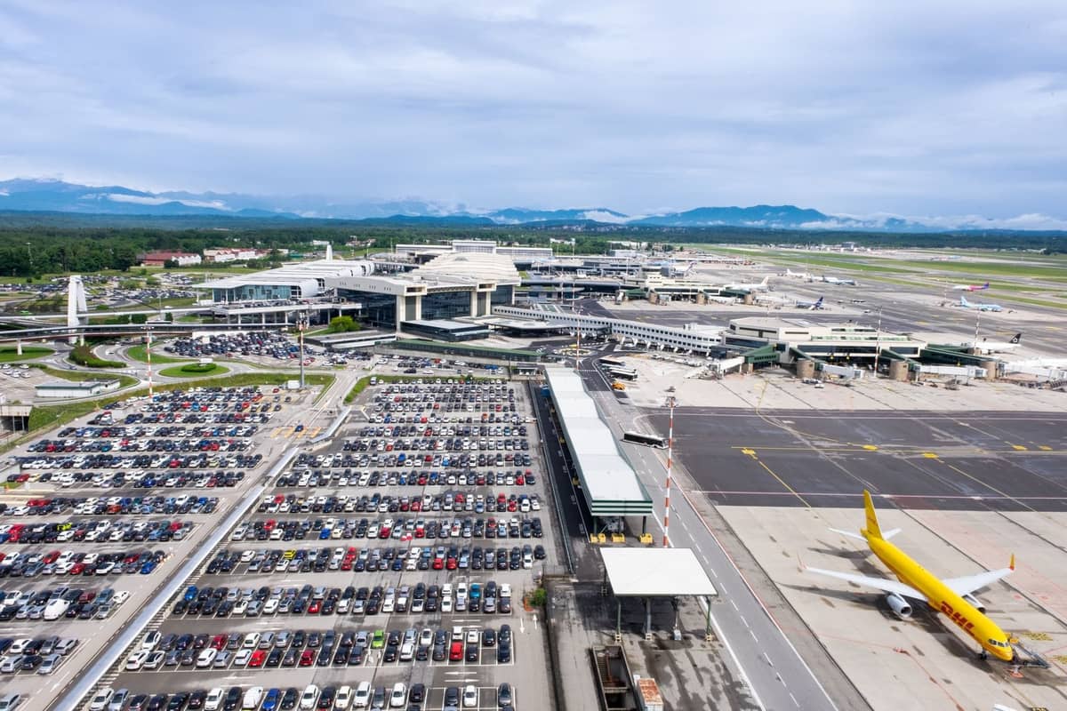 Luftaufnahme eines großen Flughafens mit mehreren Terminals, geparkten Flugzeugen und weitläufigen Parkplätzen vor der Kulisse ferner Berge.