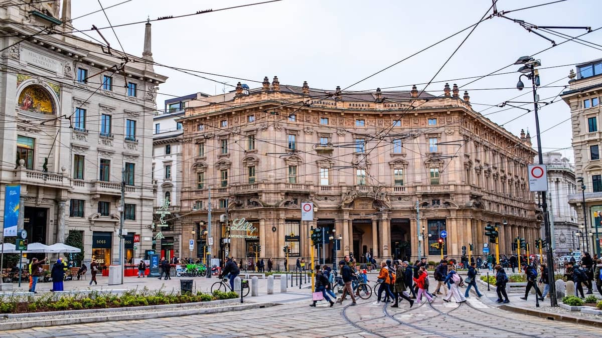 Trafficato incrocio nel centro di Milano con edifici storici, binari del tram e pedoni che attraversano la strada.