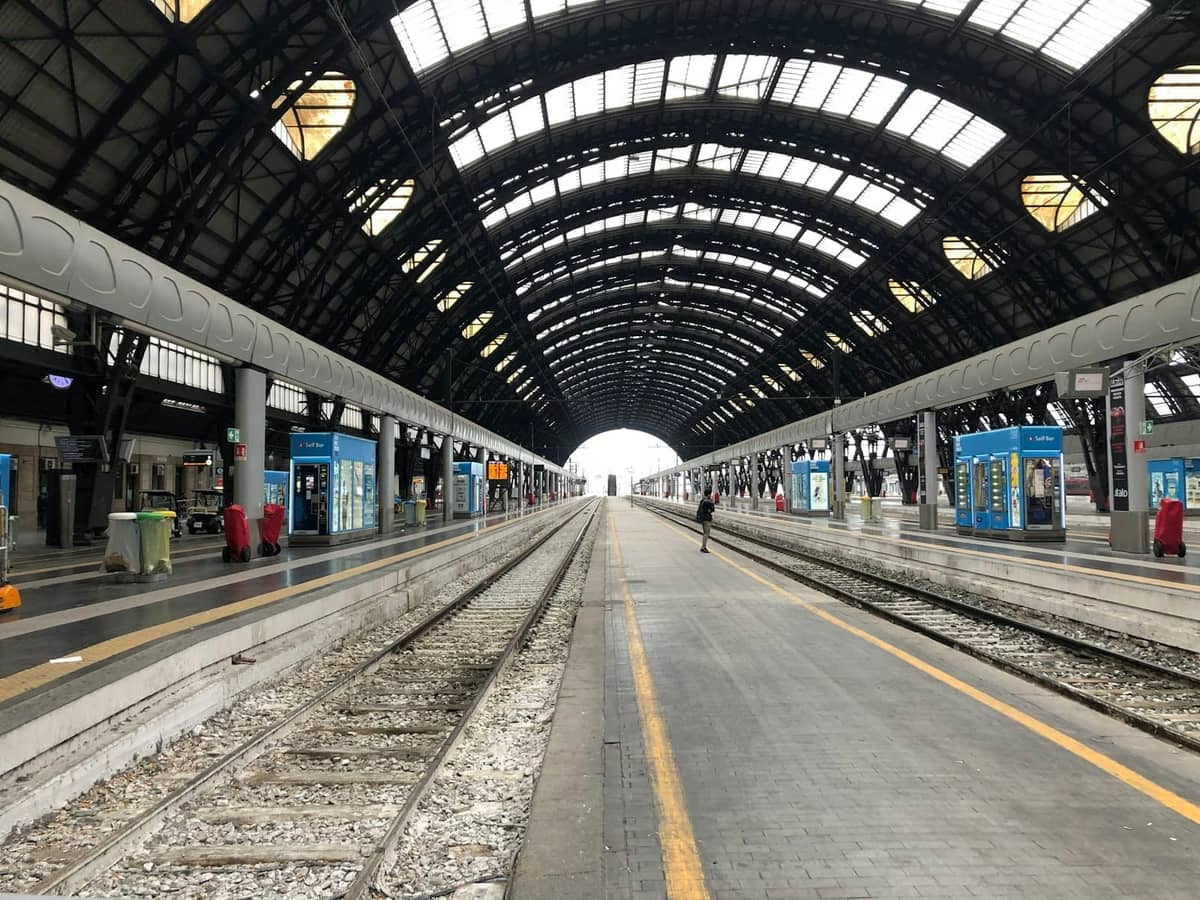 Interno della grande stazione ferroviaria di Milano con tetto ad arco in acciaio e vetro, binari vuoti e chioschi moderni