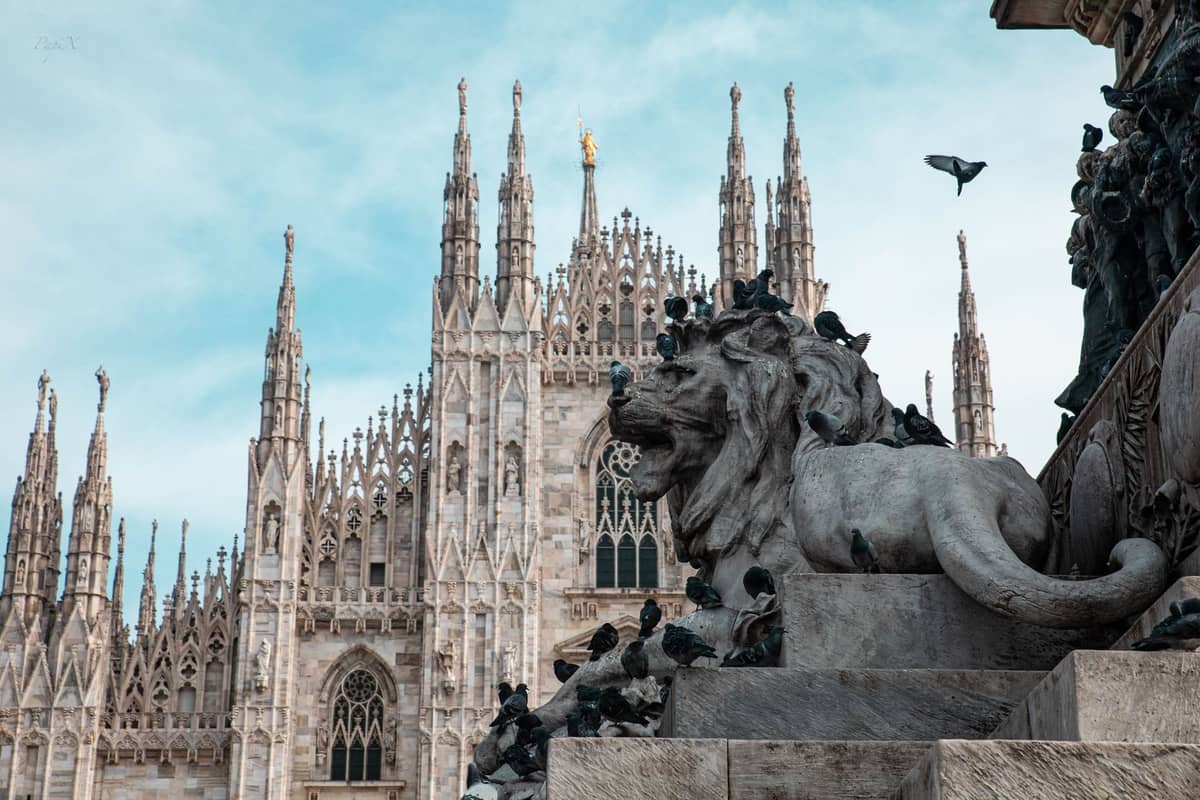 Vista della facciata del Duomo di Milano con guglie gotiche e statue, scultura di leone in pietra e piccioni in primo piano sotto un cielo azzurro.
