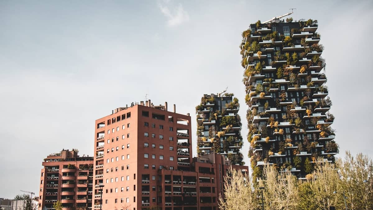 Vista ampia delle torri gemelle del Bosco Verticale di Milano ricoperte di verde, accanto a moderni edifici in mattoni rossi e alberi in primo piano in una giornata serena.