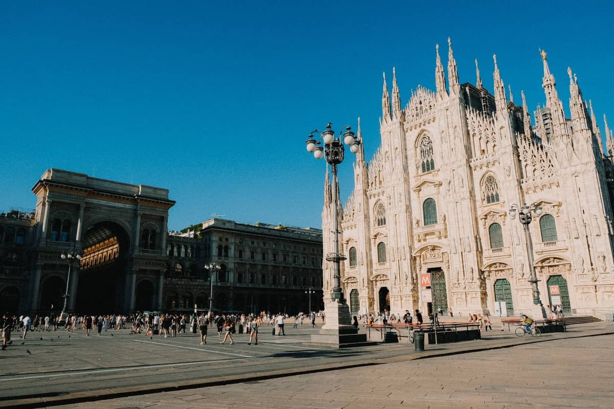 Weitwinkelansicht der Mailänder Kathedrale Duomo mit der Galleria Vittorio Emanuele II und lebhaften Menschenmengen im umliegenden Viertel unter einem klaren blauen Himmel.