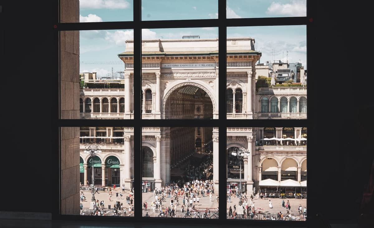 Vista attraverso una grande finestra a griglia sulla Galleria Vittorio Emanuele II di Milano, animata da persone e da un'elegante architettura storica.