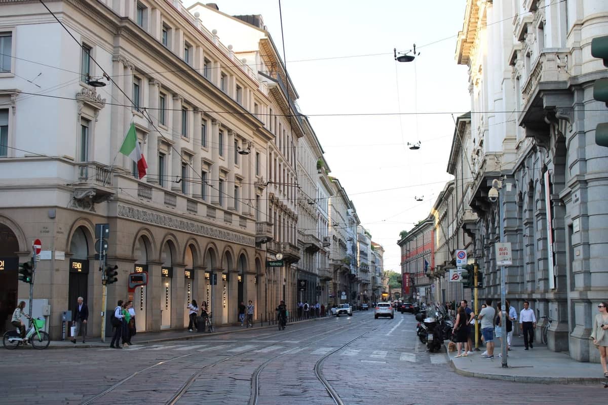 Ampia strada acciottolata a Milano fiancheggiata da eleganti edifici, bandiere italiane, binari del tram e pedoni che esplorano il centro città.