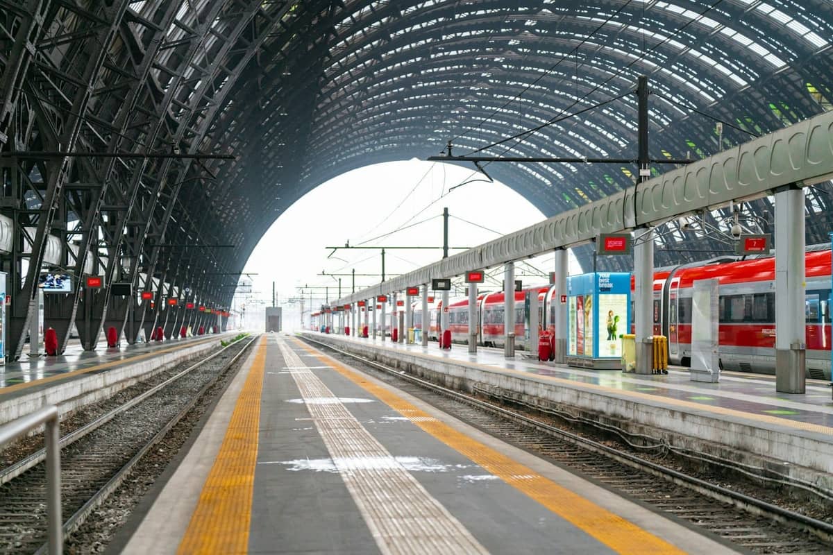Vista ampia dei binari della stazione Milano Centrale con treni rossi e tetto ad arco in vetro, un iconico snodo di viaggio per spostarsi a Milano.