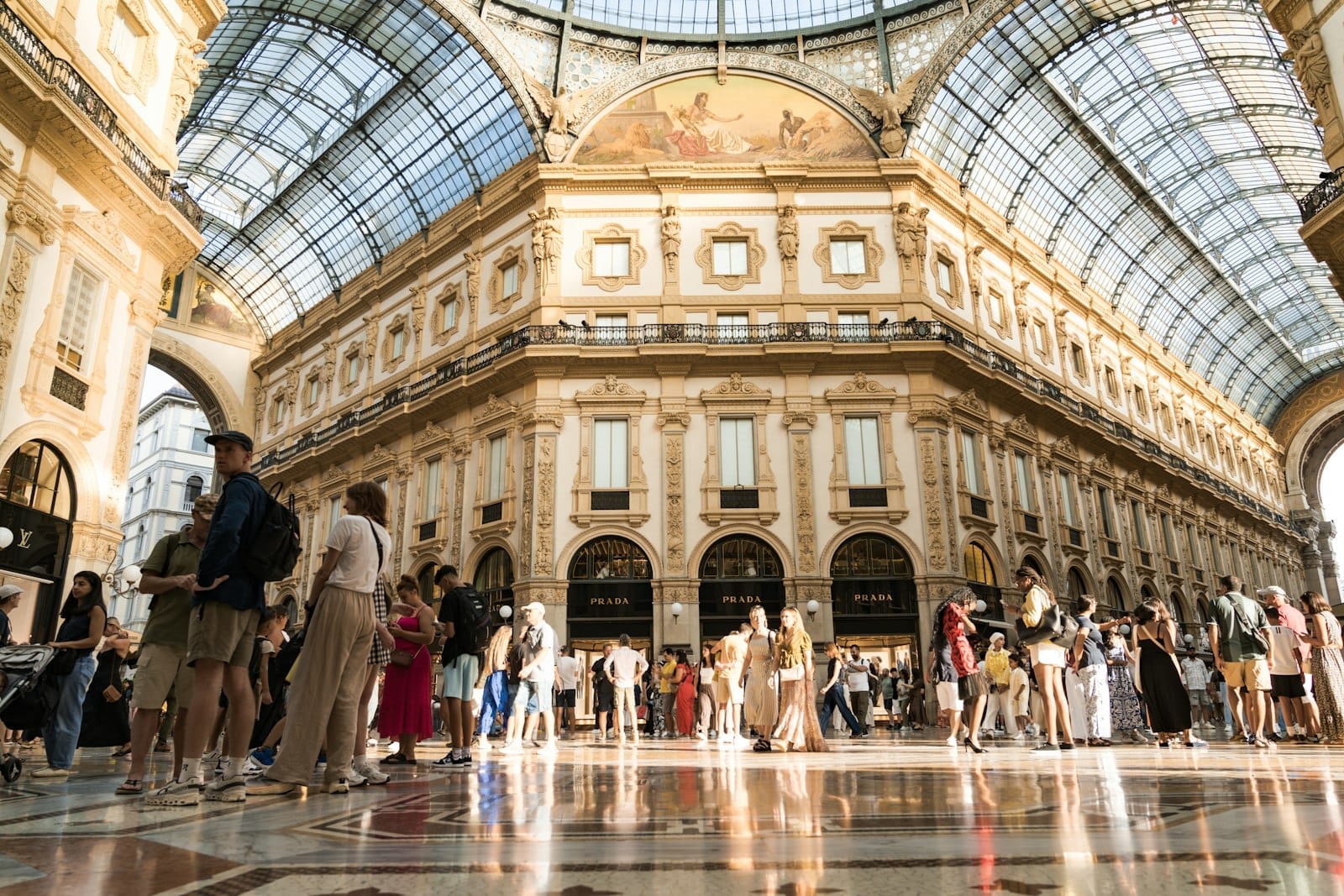 Vista ampla da Galleria Vittorio Emanuele II em Milão, com compradores elegantes sob um imenso teto de vidro em um dia ensolarado.