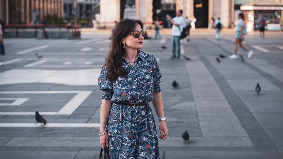 Frau in gemustertem Kleid und Sonnenbrille, die über die Piazza del Duomo in Mailand läuft, mit Tauben und klassischer Architektur im Hintergrund.