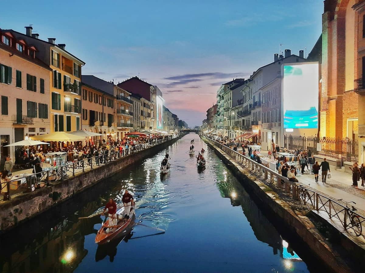 Vista nocturna del barrio Navigli de Milán junto al canal, con animados restaurantes, personas cenando al aire libre y barcas en el canal bajo un cielo de colores.
