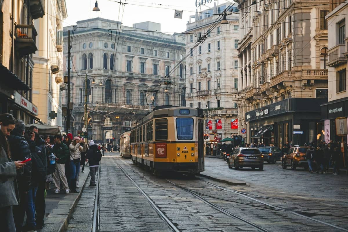 Eine klassische gelbe Straßenbahn fährt durch eine belebte Straße in Mailand, während Menschen warten und im Hintergrund prächtige historische Gebäude zu sehen sind.