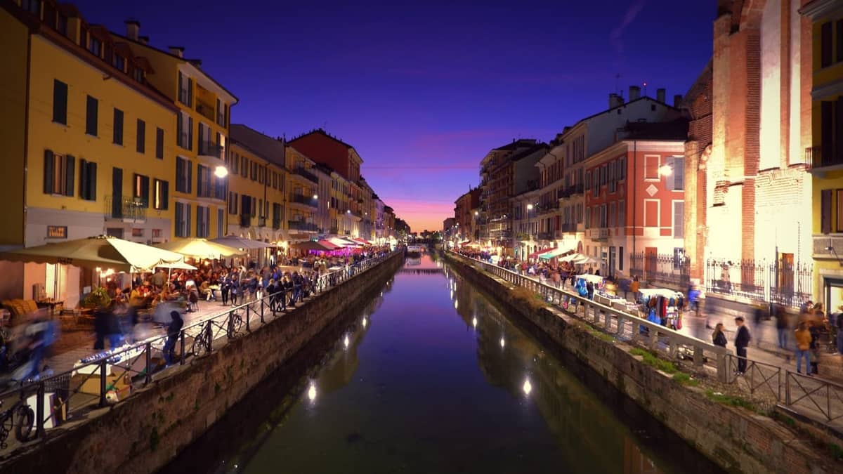 Abendlicher Blick auf den Navigli-Kanal in Mailand mit lebhaften Gebäuden, belebten Menschenmengen und Spiegelungen im Wasser bei Sonnenuntergang.