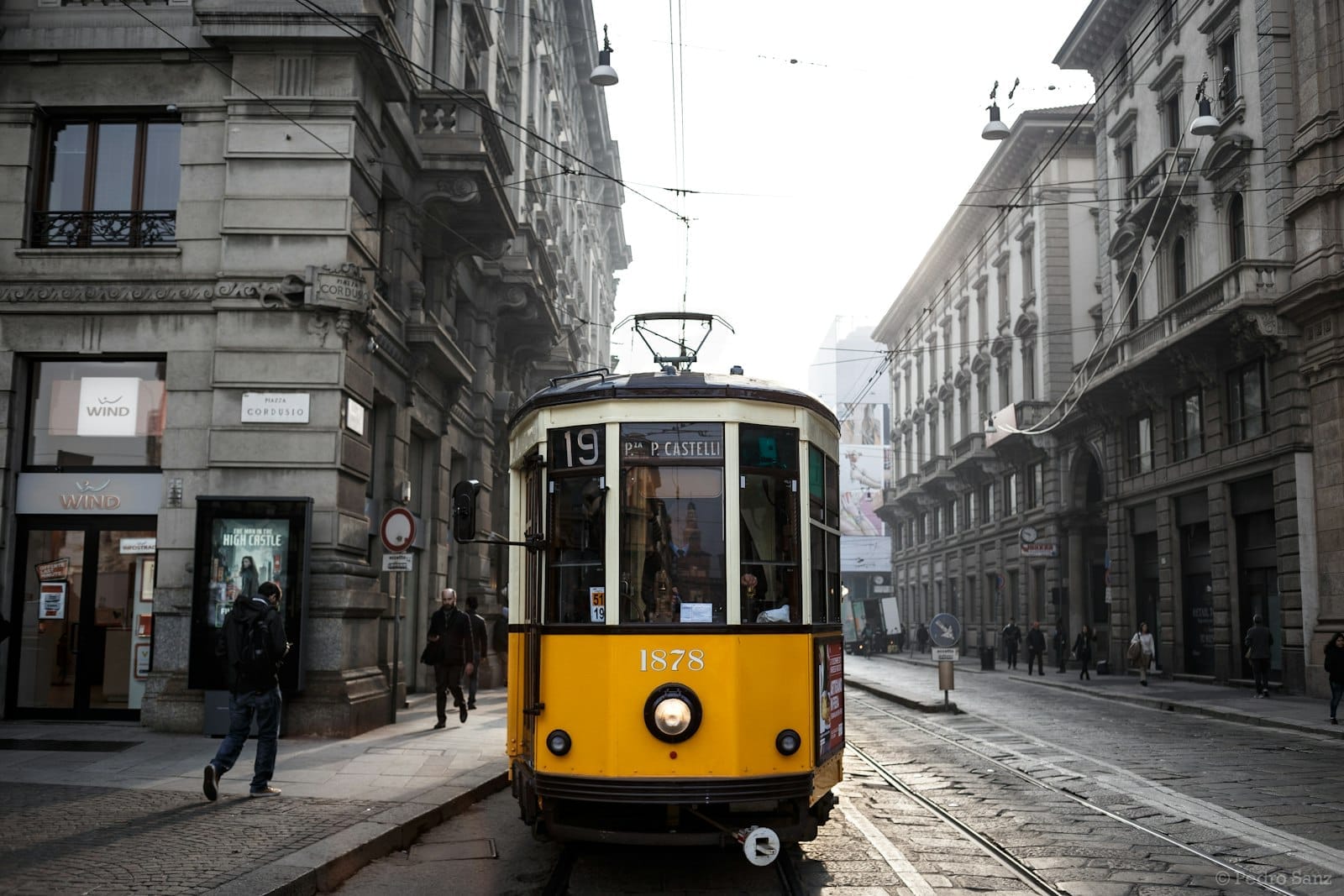 O clássico bonde amarelo número 19 percorrendo uma rua estreita ladeada por prédios históricos de pedra no centro de Milão numa manhã com neblina.