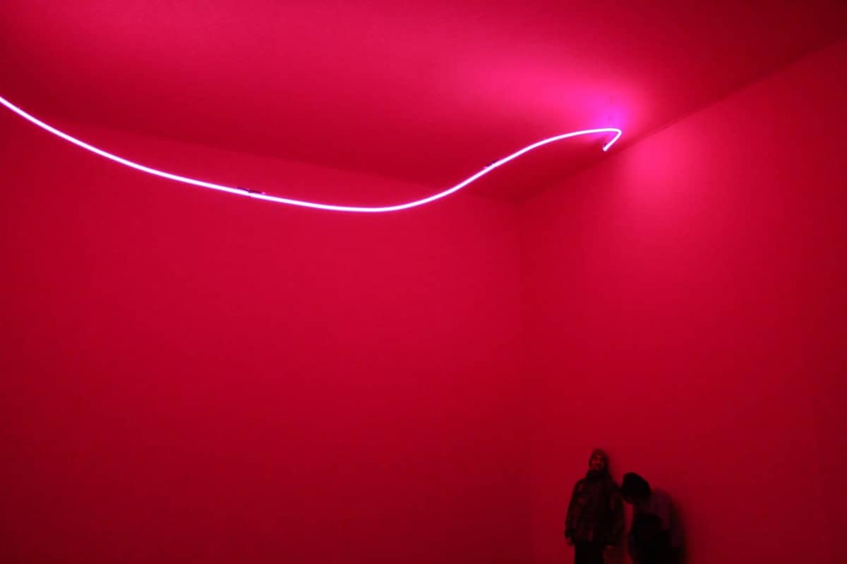 Eine zeitgenössische Kunstinstallation mit einem neonfarbenen rosa Licht in einem minimalistischen, leuchtend roten Raum, zwei Figuren stehen in der Ecke