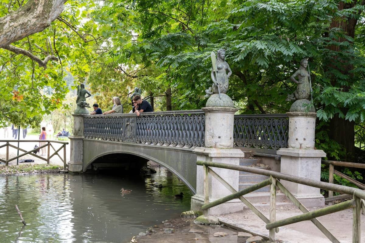 Kleine Zierbrücke über einen Teich, umgeben von üppigem Grün in einem ruhigen Park in Mailand, mit Menschen, die sich auf das Geländer stützen.