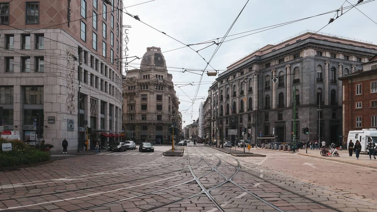 Breite Kreuzung in Mailand mit Straßenbahnschienen, großartiger Architektur aus dem frühen 20. Jahrhundert und Oberleitungen unter einem bewölkten Himmel.