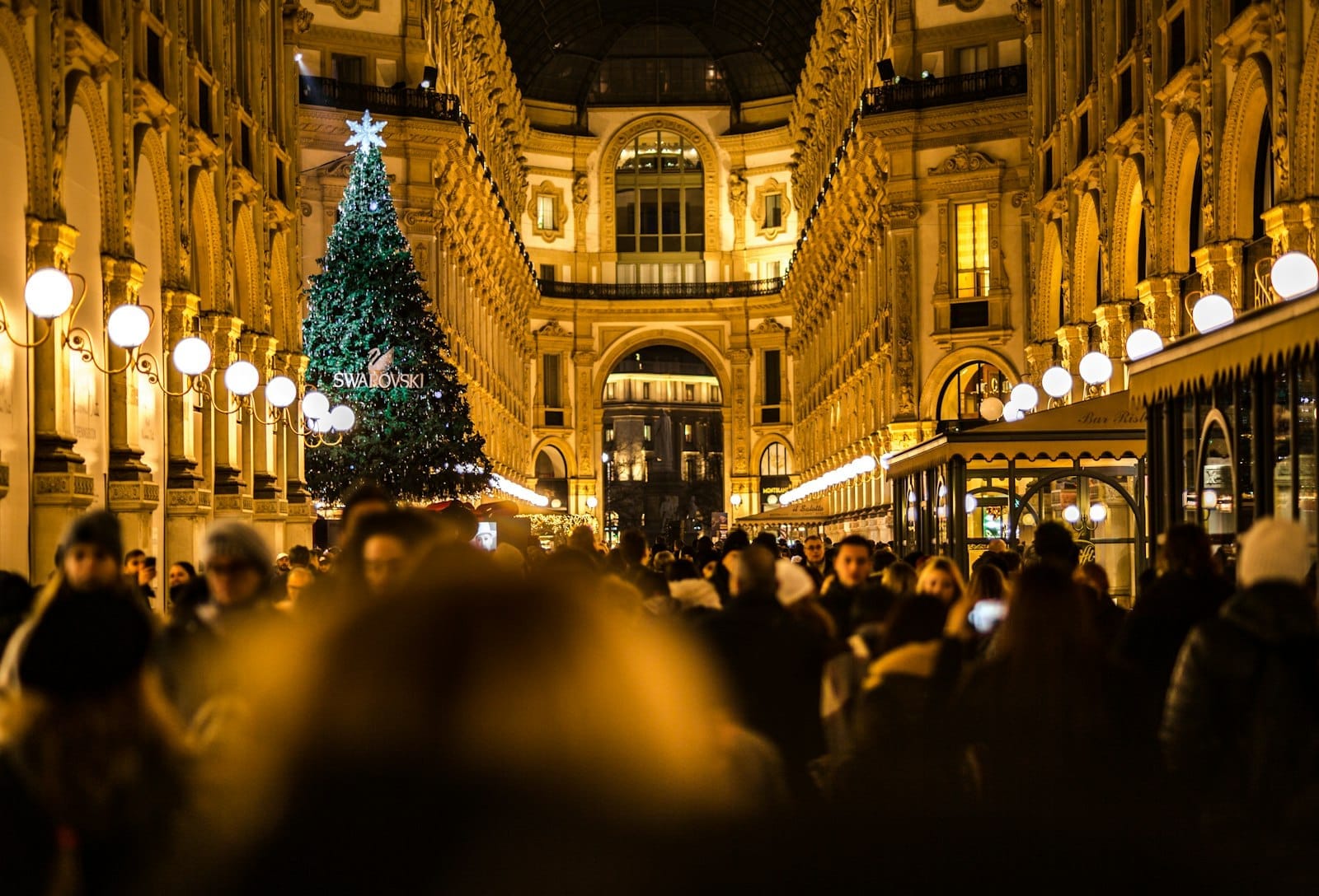 Multidão aprecia o clima festivo na Galleria Vittorio Emanuele II, em Milão, com uma grande árvore de Natal e luzes natalinas brilhando à noite.