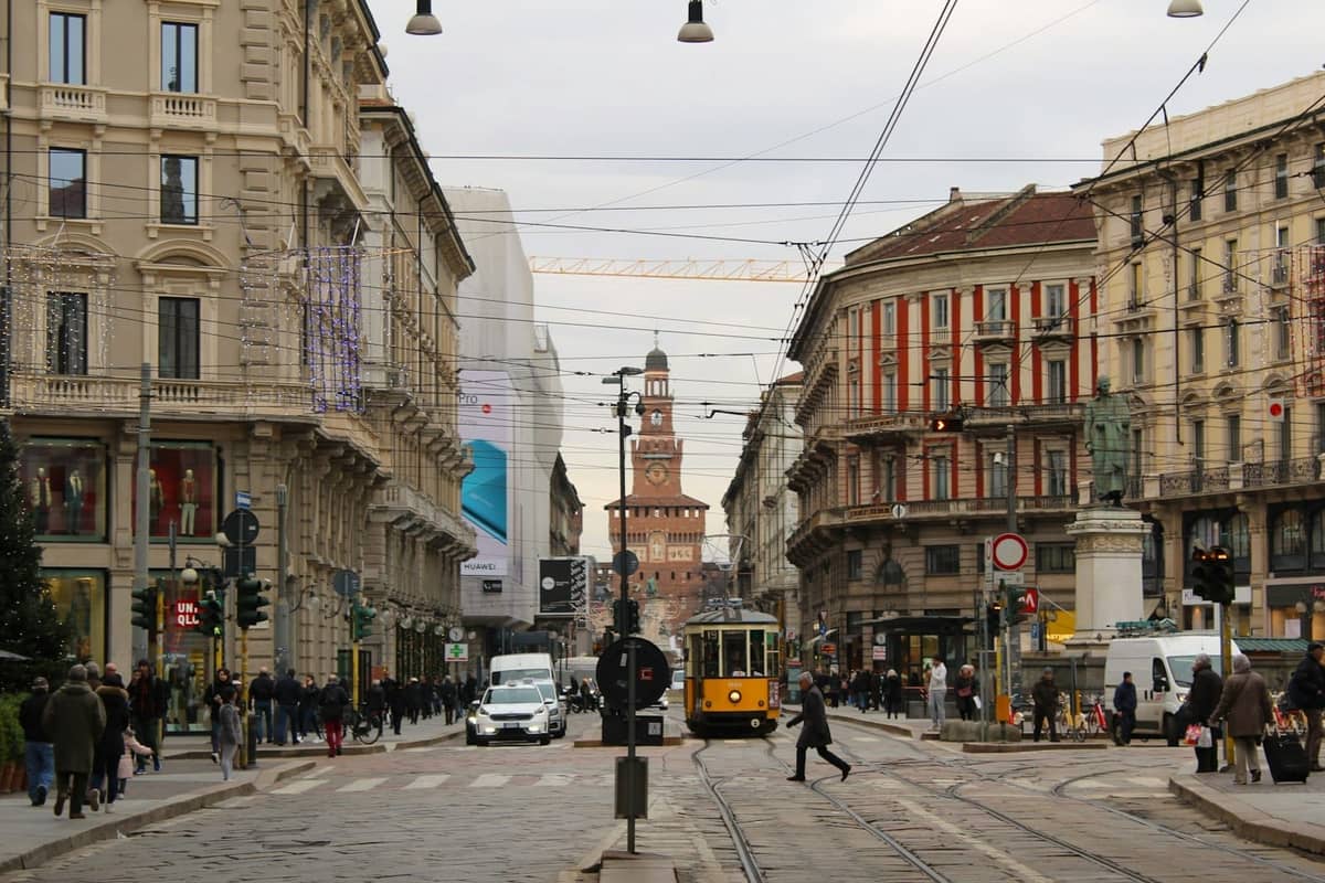 Breite Stadtstraße in Mailand mit einer gelben Straßenbahn, Fußgängern, Autos und dem Castello Sforzesco im Hintergrund.