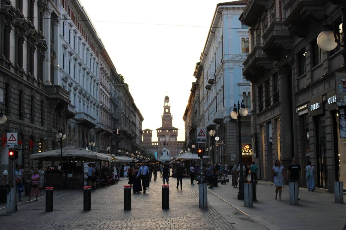 Straßenszene im Zentrum von Mailand mit Fußgängern, gesäumt von historischen Gebäuden, mit dem Castello Sforzesco im Hintergrund bei Sonnenuntergang