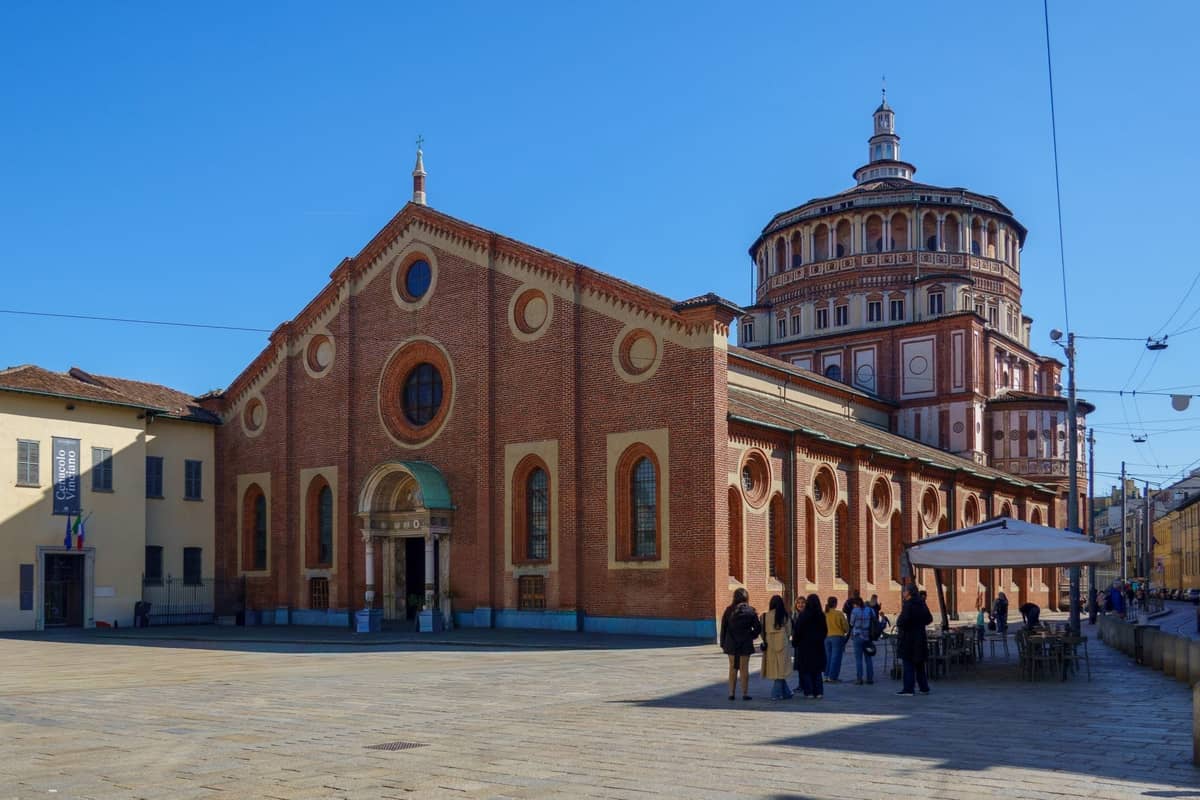 Weitwinkelansicht von Santa Maria delle Grazie in Mailand, mit der ikonischen Backsteinsfassade und Kuppel, Menschen versammeln sich an einem sonnigen Tag draußen.