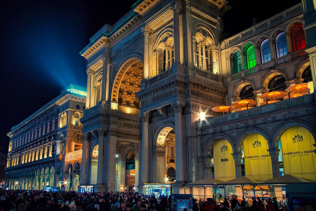 Menschenmassen an der Galleria Vittorio Emanuele II in Mailand bei Nacht beleuchtet, mit erleuchteten Gebäuden und Menschen, die sich draußen versammeln.