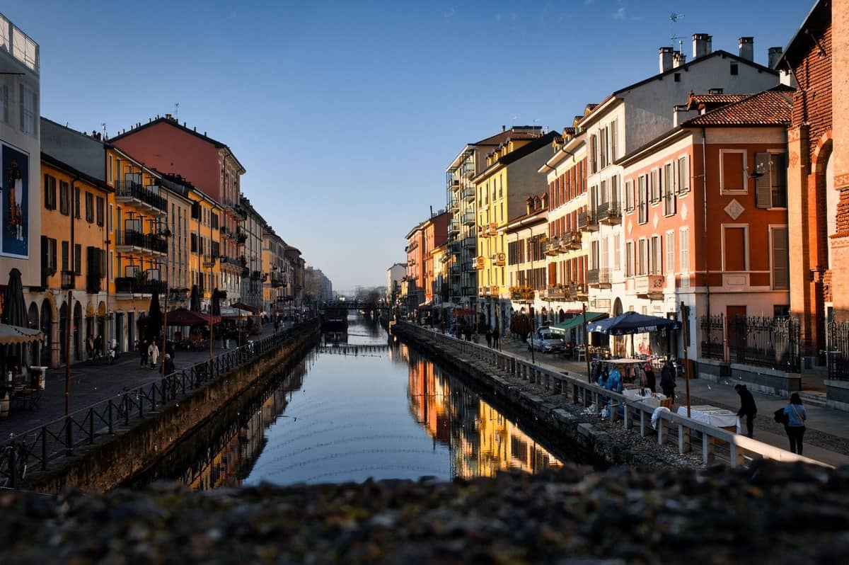Słoneczny widok na dzielnicę Navigli w Mediolanie z kolorowymi budynkami wzdłuż kanału, odbijającymi się w wodzie, i ludźmi spacerującymi po obu stronach.
