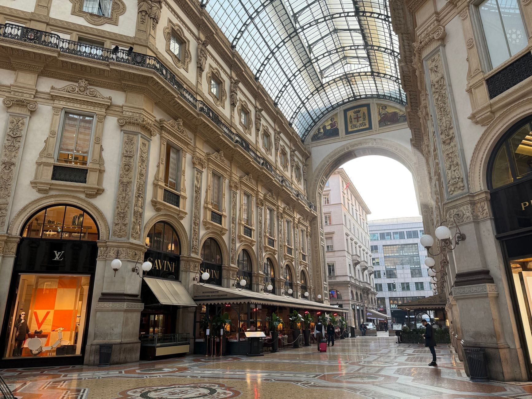 Interior elegante da Galleria Vittorio Emanuele II em Milão, com lojas de luxo, arquitetura ornamentada e luz natural entrando pelo teto de vidro.