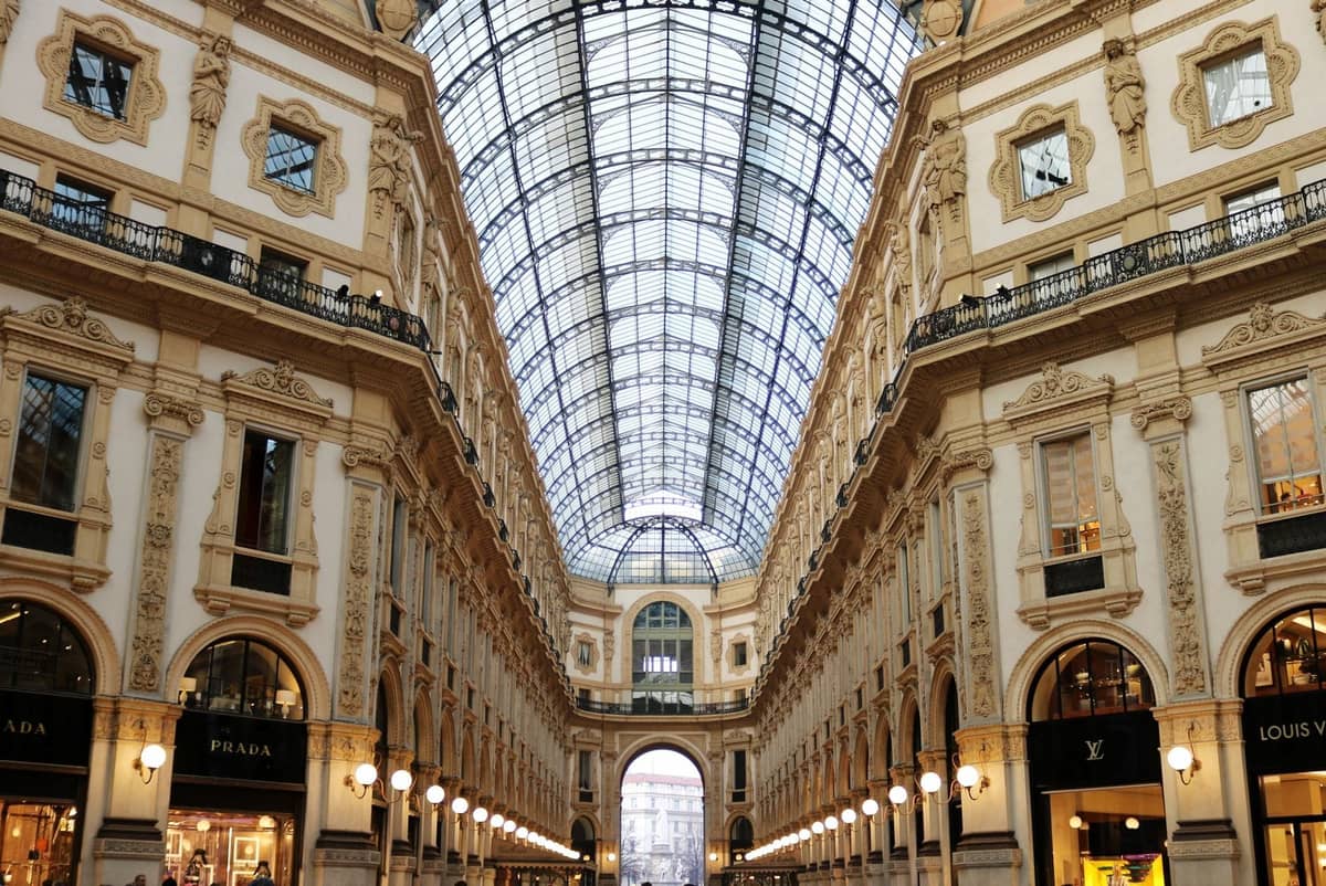 Szeroki widok wnętrza Galleria Vittorio Emanuele II w Mediolanie z ozdobnymi fasadami, szklanym sklepieniem kopułowym i szyldami luksusowych butików po obu stronach.