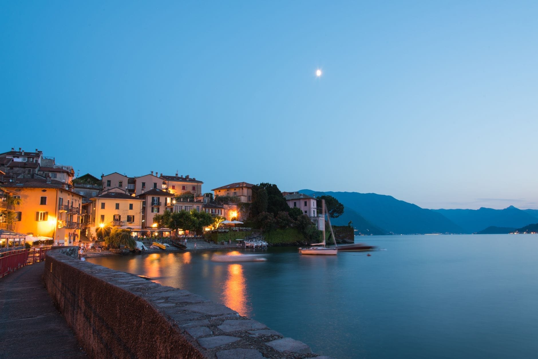 Vila à beira do Lago de Como ao entardecer com luzes refletindo na água, veleiro ancorado e montanhas ao fundo sob um céu crepuscular.