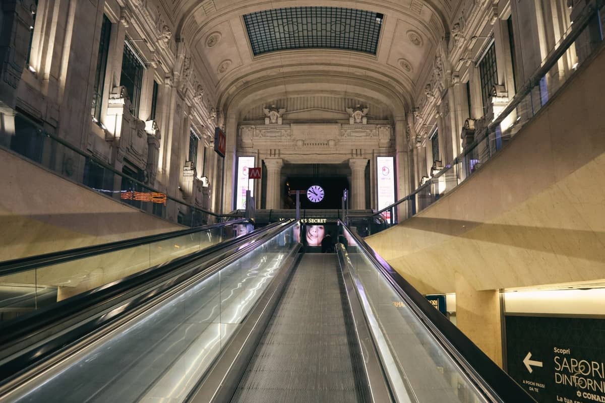 Góc nhìn từ thang cuốn bên trong ga Milano Centrale, hiện ra kiến trúc hoành tráng với các vòm cuốn, cột trụ và đồng hồ phía trên lối vào.