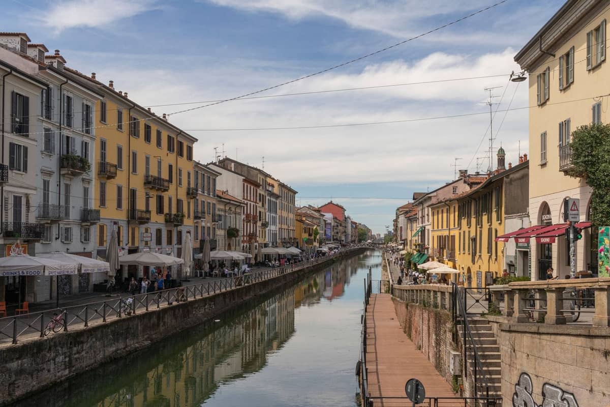 Milano'nun Navigli kanal bölgesinin gündüz görünümü; renkli binalar, açık hava kafeleri, canlı kaldırımlar ve mevsimsel etkinliği gösteren açık gökyüzüyle.
