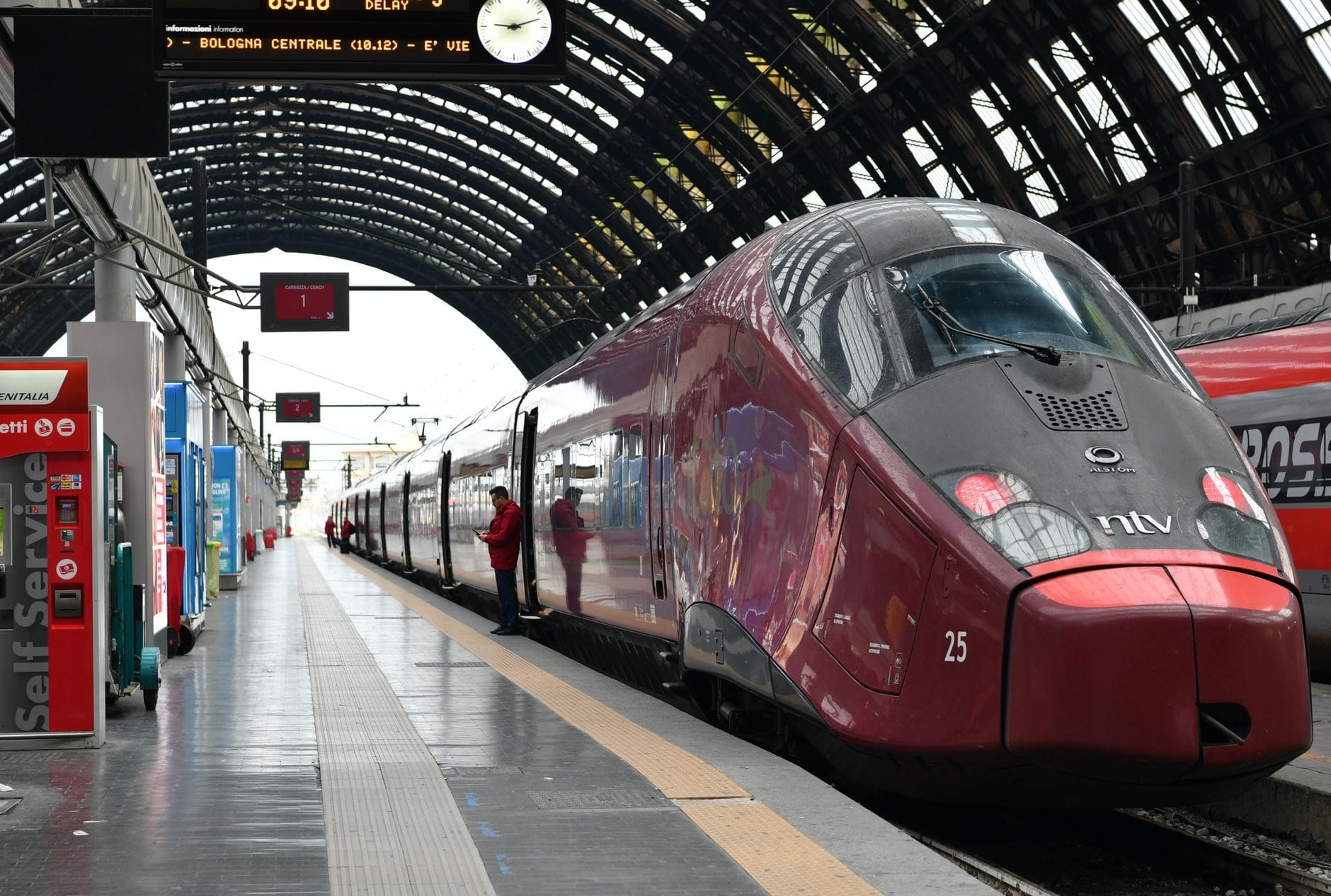 Trem vermelho de alta velocidade Italo parado na plataforma da grandiosa estação central de Milão, com um viajante ao lado da composição.