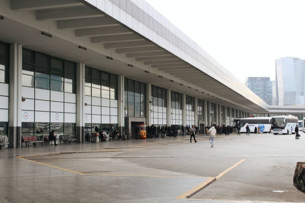 Milano'da modern bir otobüs terminali; arka planda insanlar, otobüsler ve şehir binaları, büyük bir saçak altında.