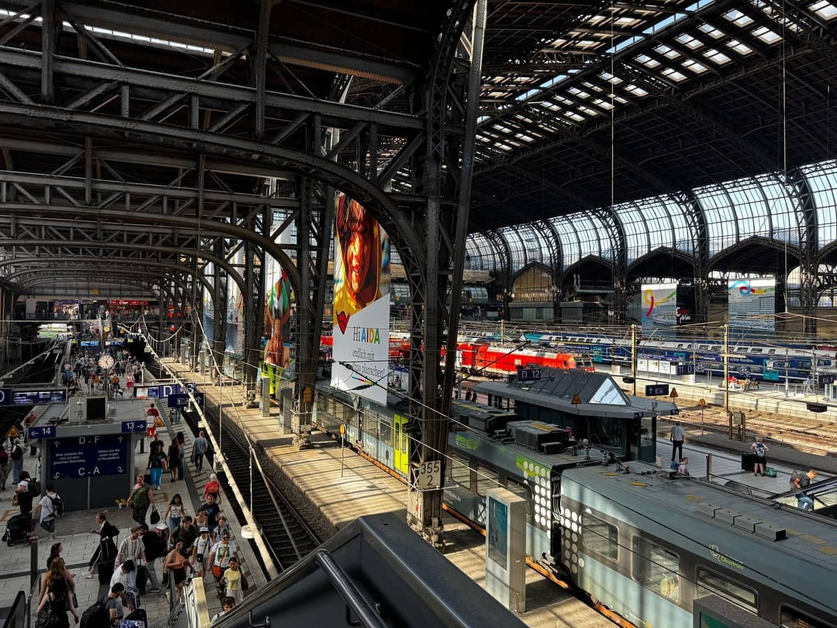 Milano Centrale tren istasyonunun içinin geniş görünümü; raylardaki trenler ve kemerli cam çatının altında platformlarda yürüyen insanlar.