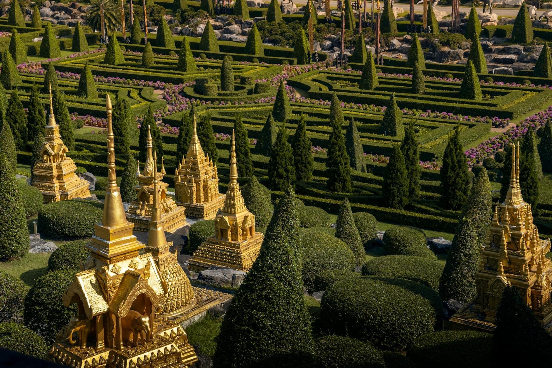 Templi thailandesi dorati in miniatura disposti tra siepi curate e alberi topiari, visti dall'alto in un parco all'aperto illuminato dal sole.