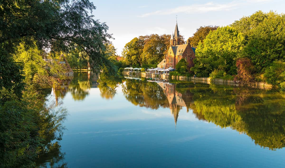 Golden hour view of Minnewater (Lake of Love) in Bruges, with the iconic brick castle-style building, lush green trees, and beautiful water reflection.