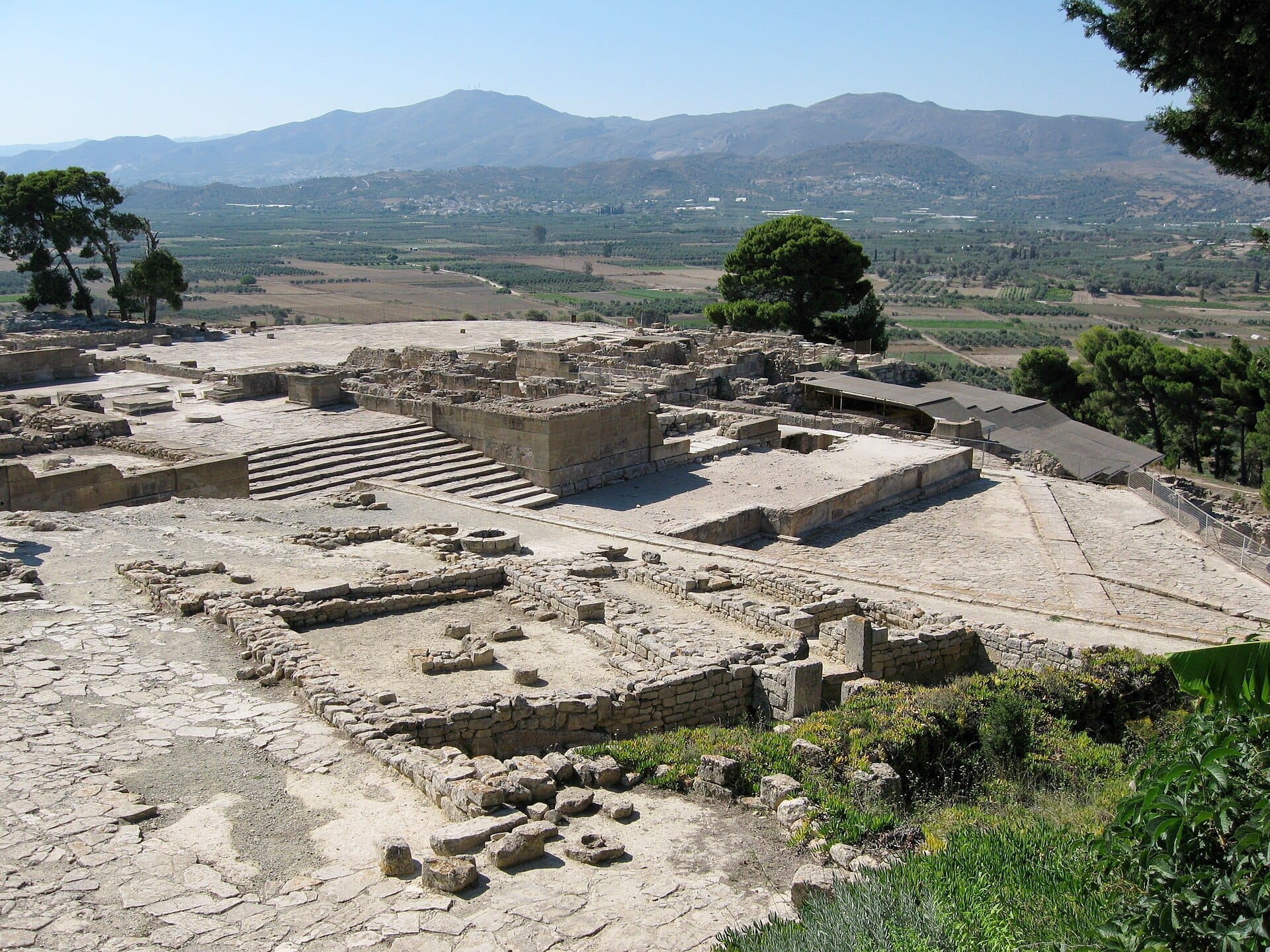 Le rovine del Palazzo di Festos con fondamenta in pietra e scalinate dominano la pittoresca pianura di Mesara e le lontane montagne di Creta sotto un cielo sereno.
